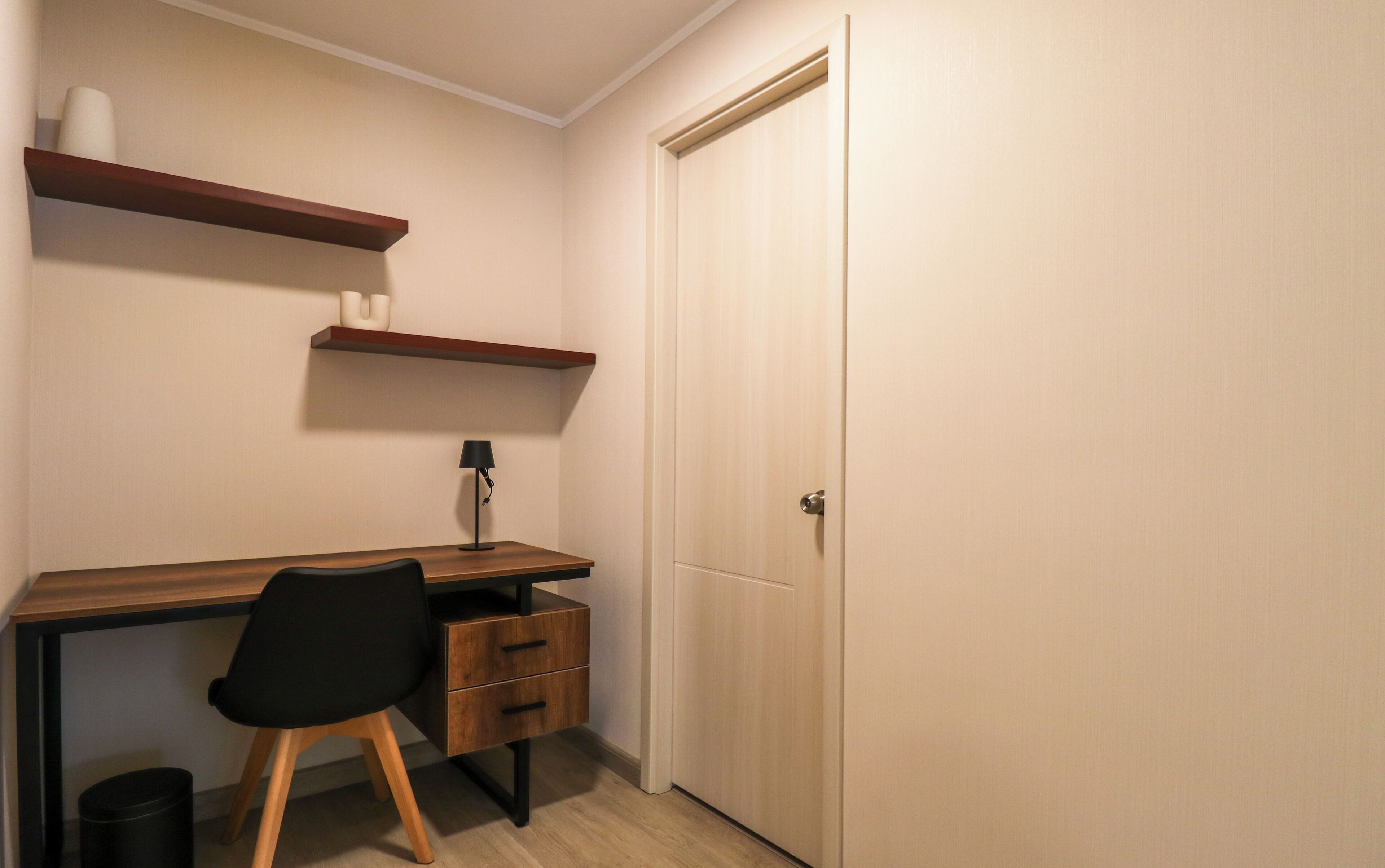 Une petite pièce, bien rangée, avec un bureau en bois, une chaise noire, une lampe et une porte fermée.