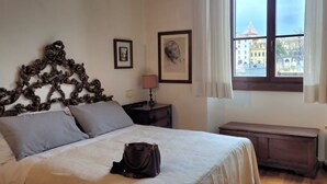 Un dormitori amb un llit gran, un panell de capçal de fusta, una taula de nit amb una làmpada, un banc i una finestra amb vista a edificis.