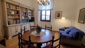 Una zona de menjador amb una taula rodona, cadires, un lampadari, una prestatgeria, una finestra i un sofà.