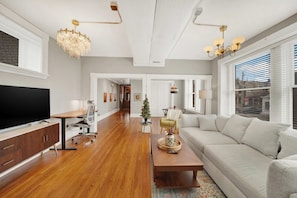 A spacious living room with a large flat-screen TV, a wooden coffee table, a grey sofa, and a chandelier. - Huge 1st floor Historic Apartment Soaring Ceilings (Wilmington)
