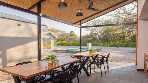 Un espace extérieur aménagé pour les repas, avec des tables en bois et des chaises noires, un ventilateur de plafond, et une vue sur une piscine et un espace verdoyant.