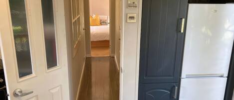 A narrow hallway with a door leading to a bedroom. The bedroom has a bed with a white comforter and a yellow pillow. There is a white refrigerator next to the door.
