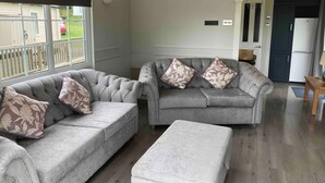 Ein Wohnzimmer mit zwei grauen Sofas, jedes mit floralen Kissen, einem kleinen Holztisch und Blick auf ein Haus durch große Fenster.