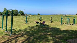 Ein Outdoor-Fitnessbereich mit Trainingsgeräten, einer Bank und Blick auf das Meer und die Hügel im Hintergrund.