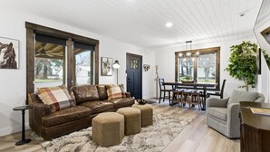 Ein geräumiges Wohnzimmer mit einem braunen Ledersofa, Ottomanen, einem grauen Sessel und einem Essbereich mit Tisch und Stühlen.