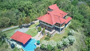 Vista aérea de una casa grande de dos pisos con techo rojo, rodeada de exuberante vegetación y una piscina.