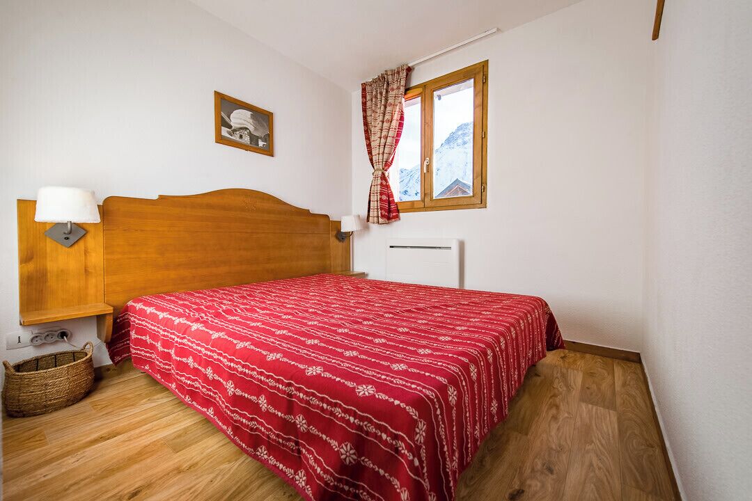 Um quarto com cabeceira de madeira, colcha de cama estampada em vermelho, luminária de cabeceira, cesta de vime e janela com vista para montanhas.