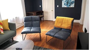 Une pièce avec un mur bleu foncé, une porte blanche et une œuvre d’art encadrée. On y trouve deux chaises, dont une recouverte d’un plaid jaune, et un canapé vert.