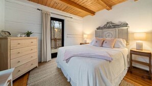 Un dormitorio con techo de madera, cama con ropa blanca, cómoda y mesita de noche con lámpara.