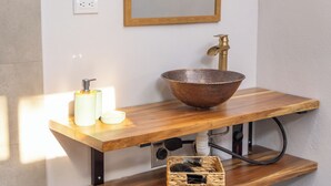 A bathroom with a wooden vanity, a copper bowl sink, a mirror, and a small shelf with a basket. - LIV 5 - Studio apartment. Minutes away from the best beaches of Guanacaste. (Lorena)