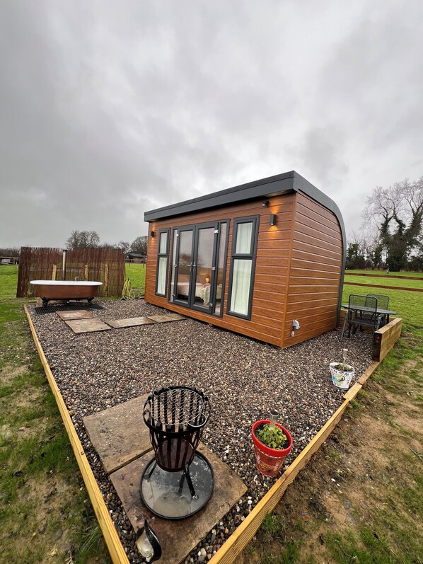 A modern wooden cabin with large windows, a patio area with a chair and a small table, and a hot tub. - Cosy off-grid countryside cabin. (Lower Boddington)
