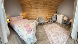 Een gezellige slaapkamer met een gebogen houten plafond, een bed met bloemenbeddels, twee stoelen, een klein bureau en een witte kast.
