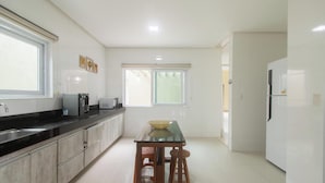 A modern kitchen with a black countertop, stainless steel sink, and white cabinets. There is a window with blinds, a refrigerator, and a table with stools. - House w/ Pool and Barbecue Grill Thg0006 (Porto Seguro)