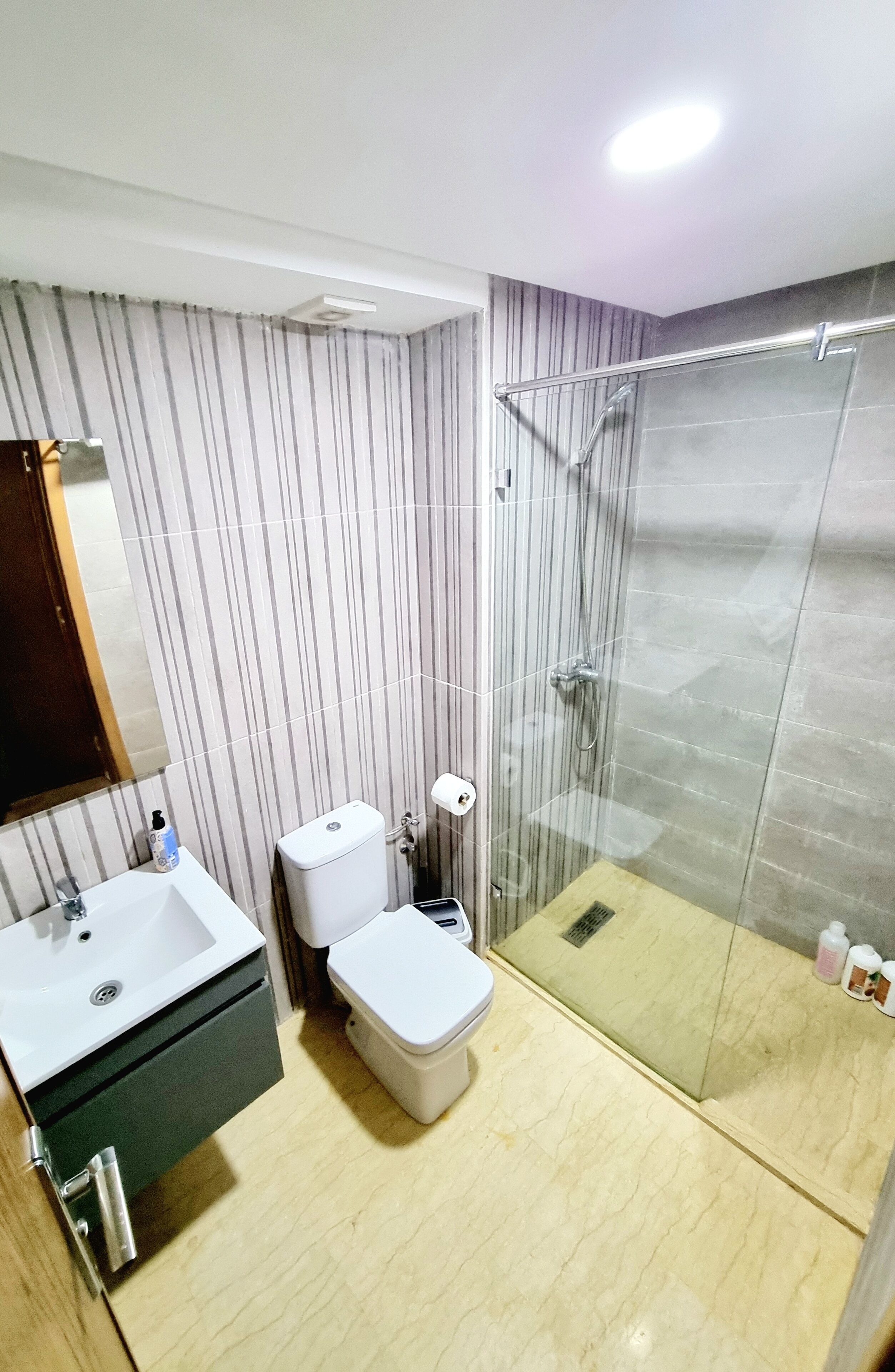 Un baño moderno con pared a rayas, cabina de ducha de vidrio, lavamanos blanco y retrete.