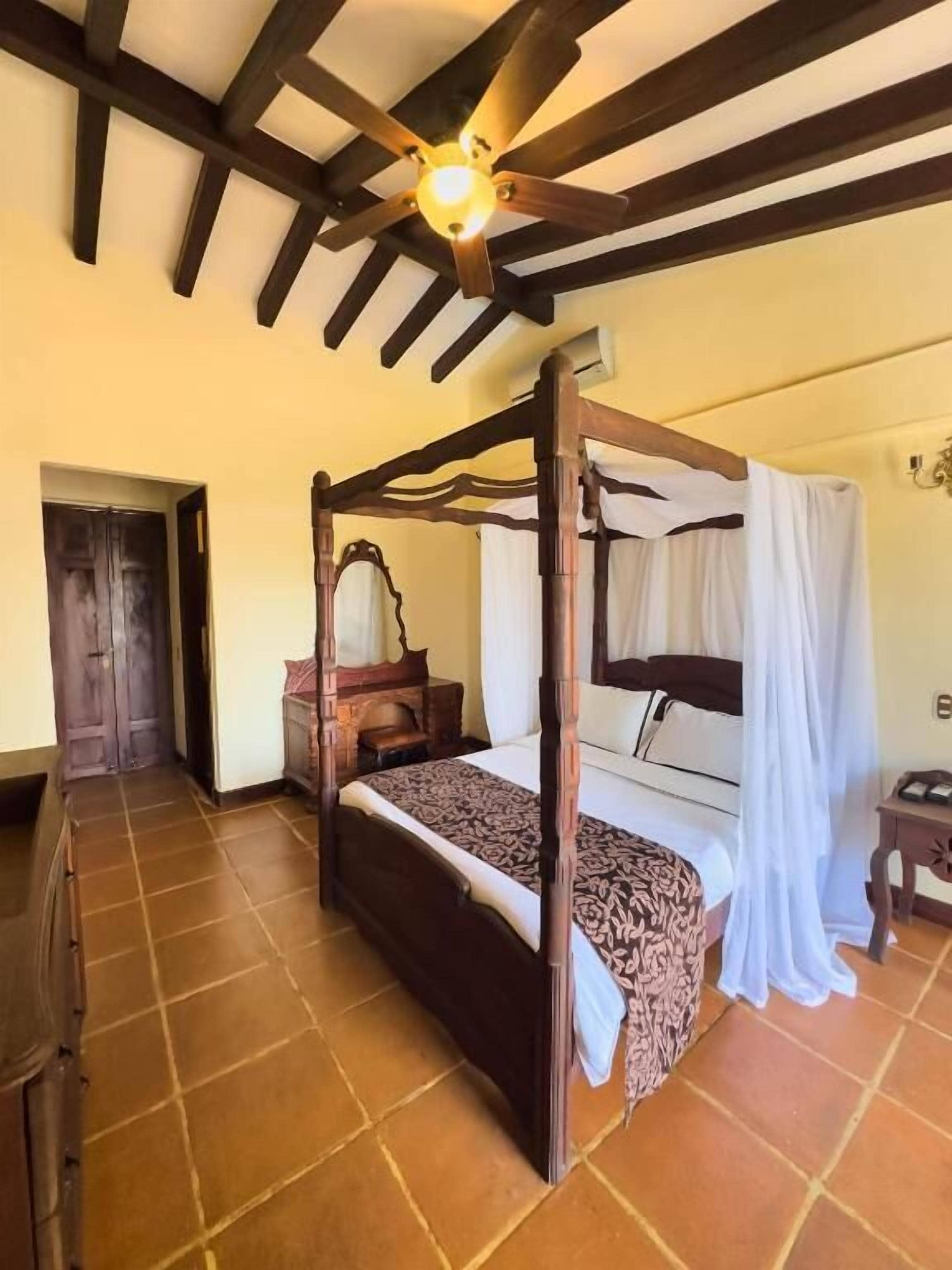 A four-poster bed with a canopy, a wooden dresser, and a ceiling fan in a room with tiled flooring.
