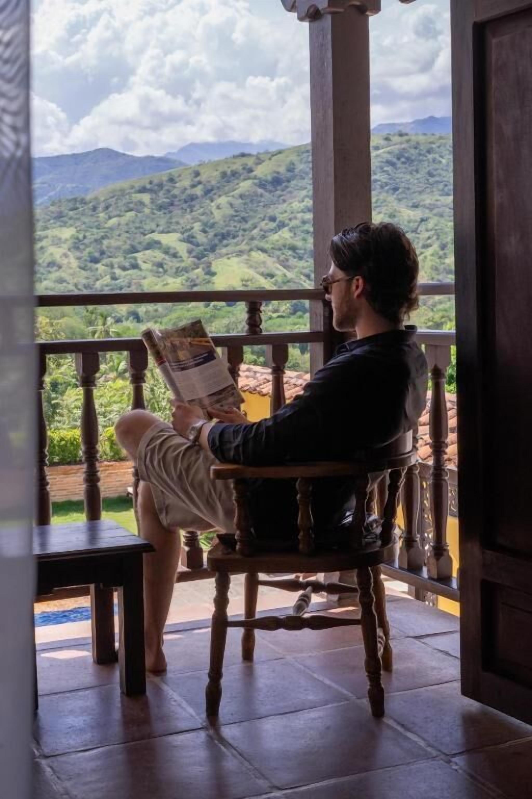 Uma pessoa sentada numa cadeira de madeira, lendo um livro numa varanda com vista para colinas verdes.