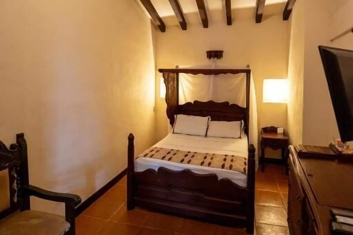 Ein Schlafzimmer mit einem hölzernen Himmelbett, einem Sessel, einem Schminktisch und einem Deckenventilator.