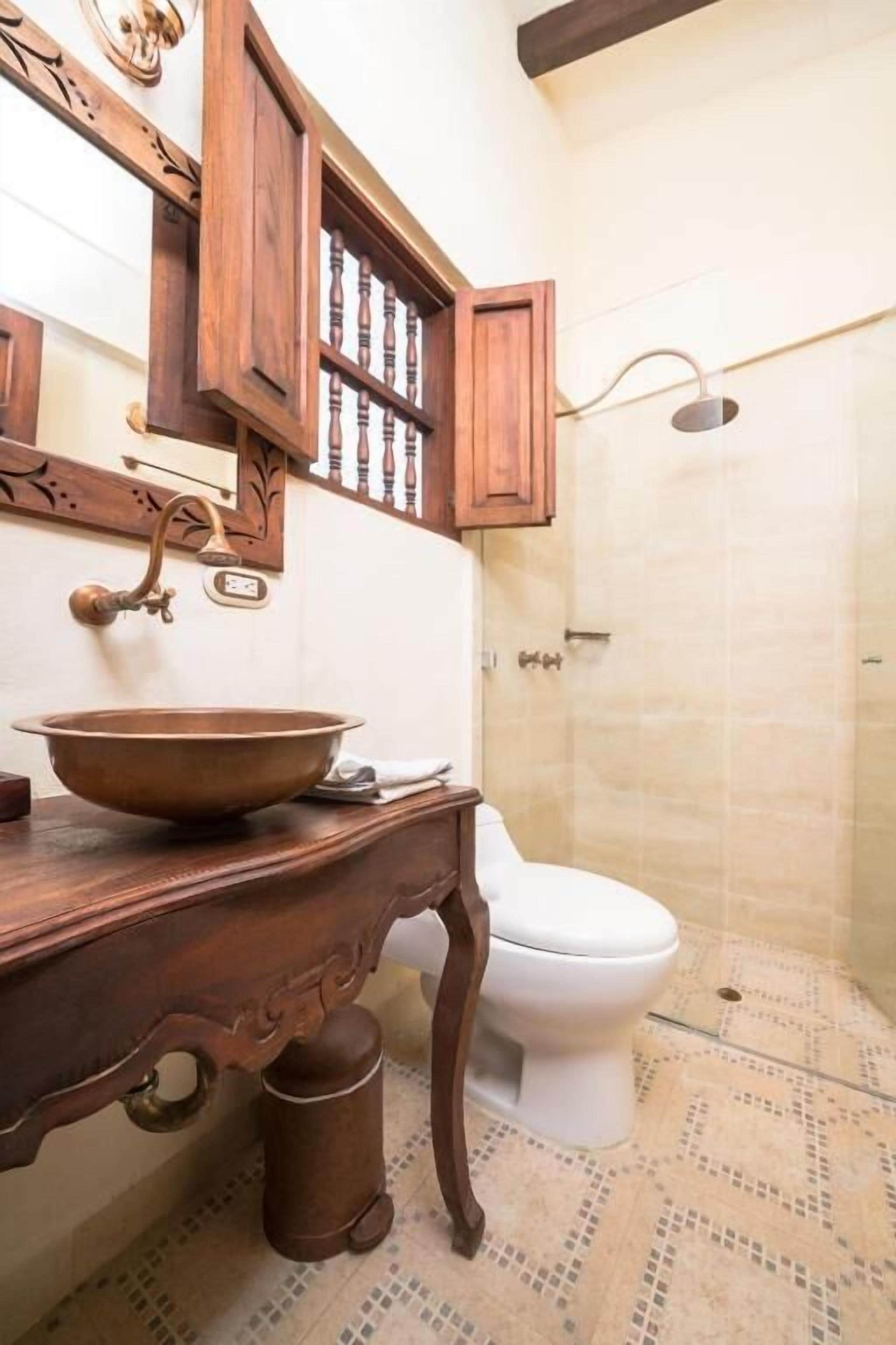 Un baño con tocador de madera, lavabo de cobre, inodoro blanco y zona de ducha.