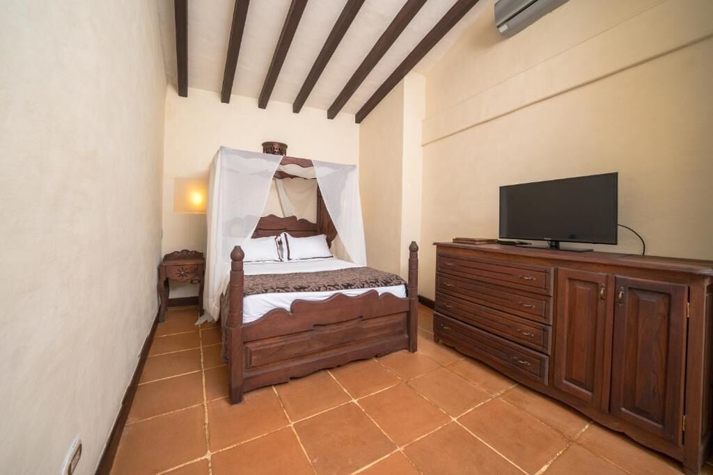 Une chambre à coucher avec un lit en bois, un téléviseur posé sur une commode en bois et une table de chevet avec une lampe.