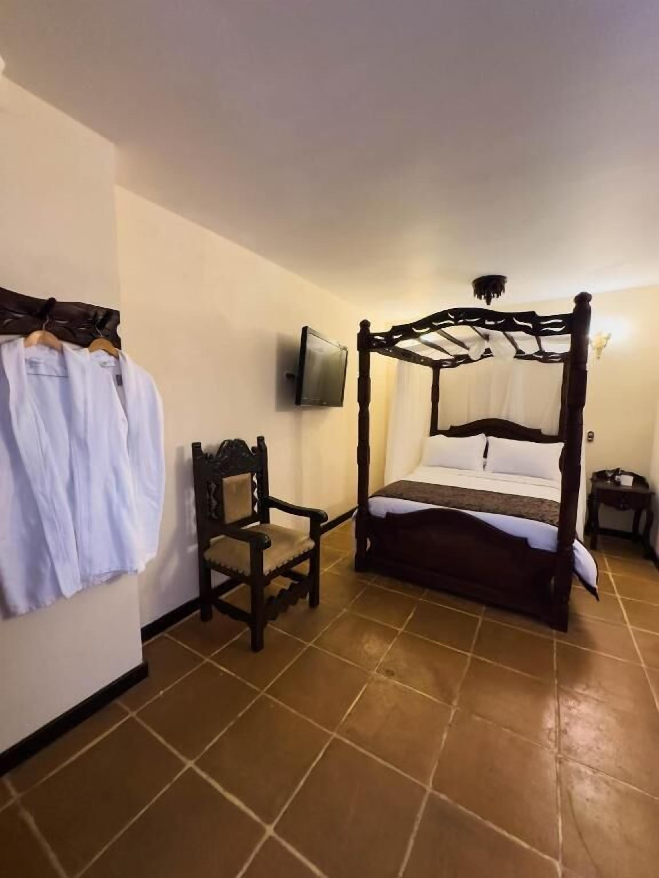 Una habitación con cama con dosel, una silla, un televisor montado en la pared y un perchero con una prenda blanca.