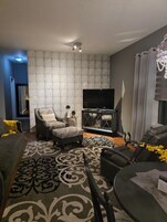 A modern living room with a grey sofa, a patterned rug, a black leather chair, a small table, and a TV.