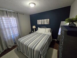 Un dormitorio con cama, mesita de noche, lámpara, cómoda y un cuadro en la pared.