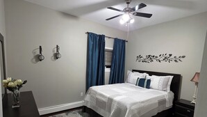 Un dormitorio con cama, ventilador de techo, mesita de noche, cómoda y un adorno en la pared.