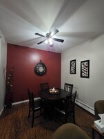 A dining room with a round table, four chairs, a ceiling fan, a wall clock, and two framed wall decorations.