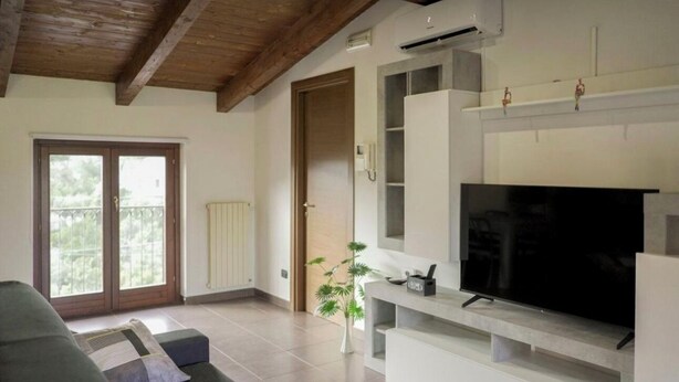 A modern living room with a large flat-screen TV, a sofa, and a sliding glass door leading to a balcony. - Casa Soele your home away from home (Pontecagnano)