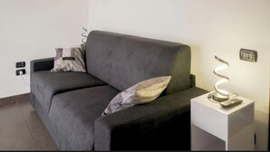 A modern living room with a dark grey sofa, two pillows, a white side table with a spiral light, and a wall socket. - Casa Soele your home away from home (Pontecagnano)