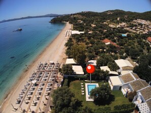 Luftaufnahme einer Küstenlandschaft mit Strand, einer Reihe von Liegestühlen und einem Schwimmbad, das von Gebäuden umgeben ist.