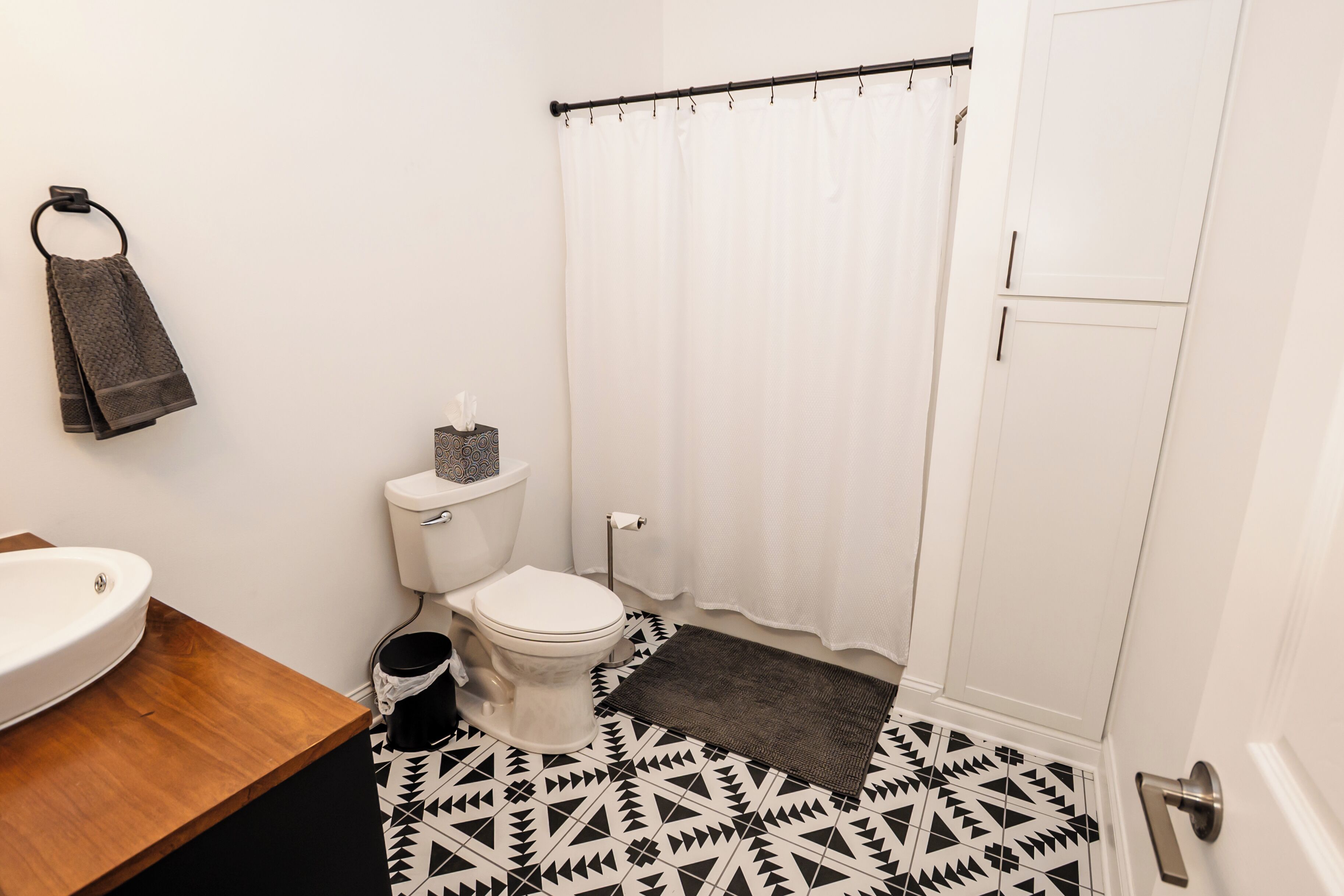 A bathroom with a white toilet, a wooden vanity with a sink, a black trash bin, a white shower curtain, and a patterned floor.