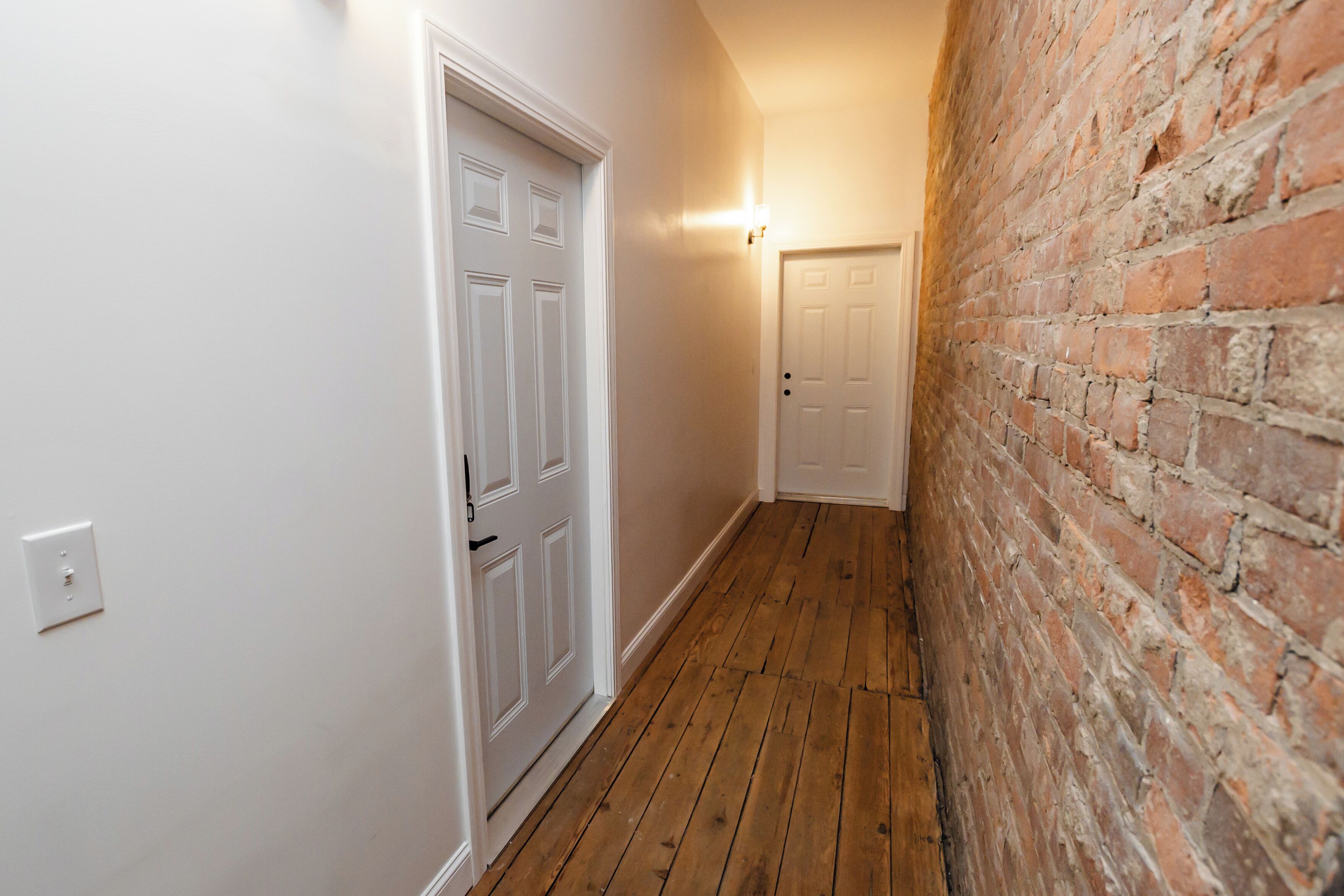A narrow hallway with wooden flooring, white walls, and a brick wall on one side.