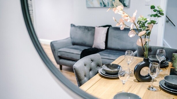 Een eethoek met een ronde tafel voor vier personen, omringd door grijze fauteuils. Op de tafel staat een vaas met roze bloemen en een zwart, rond object, mogelijk een decoratie.