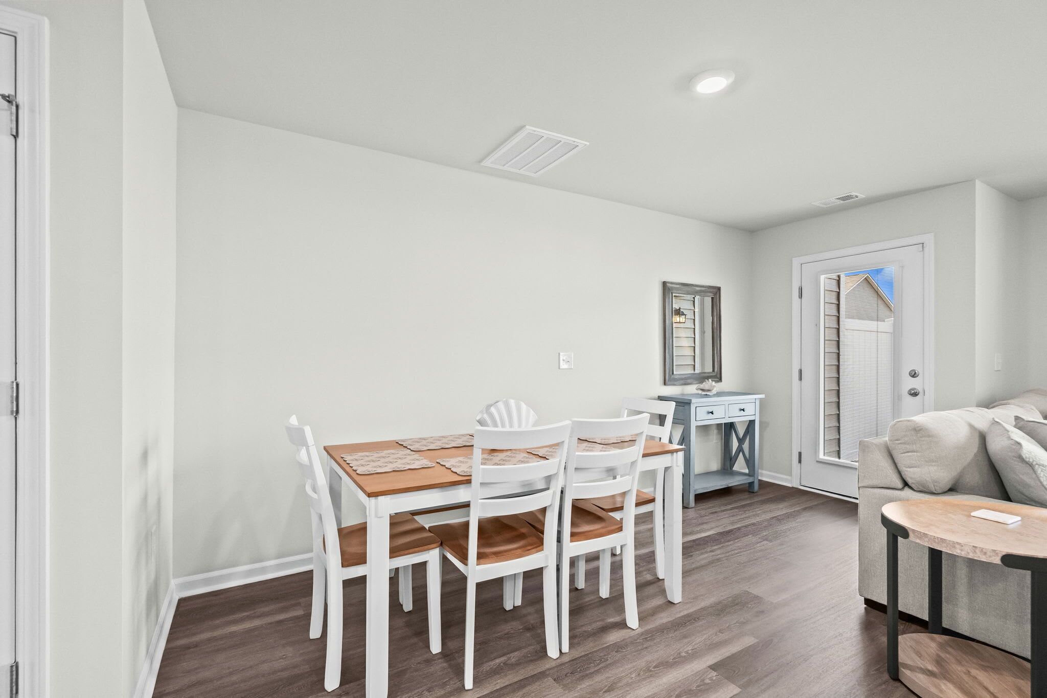 Un comedor con una mesa de madera y sillas blancas, una pequeña cocina americana con un fregadero y encimera, y una puerta que da a un balcón.