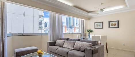 Ein modernes Wohnzimmer mit einer Couch, einem Glastisch und Blick auf die Stadt durch große Fenster.