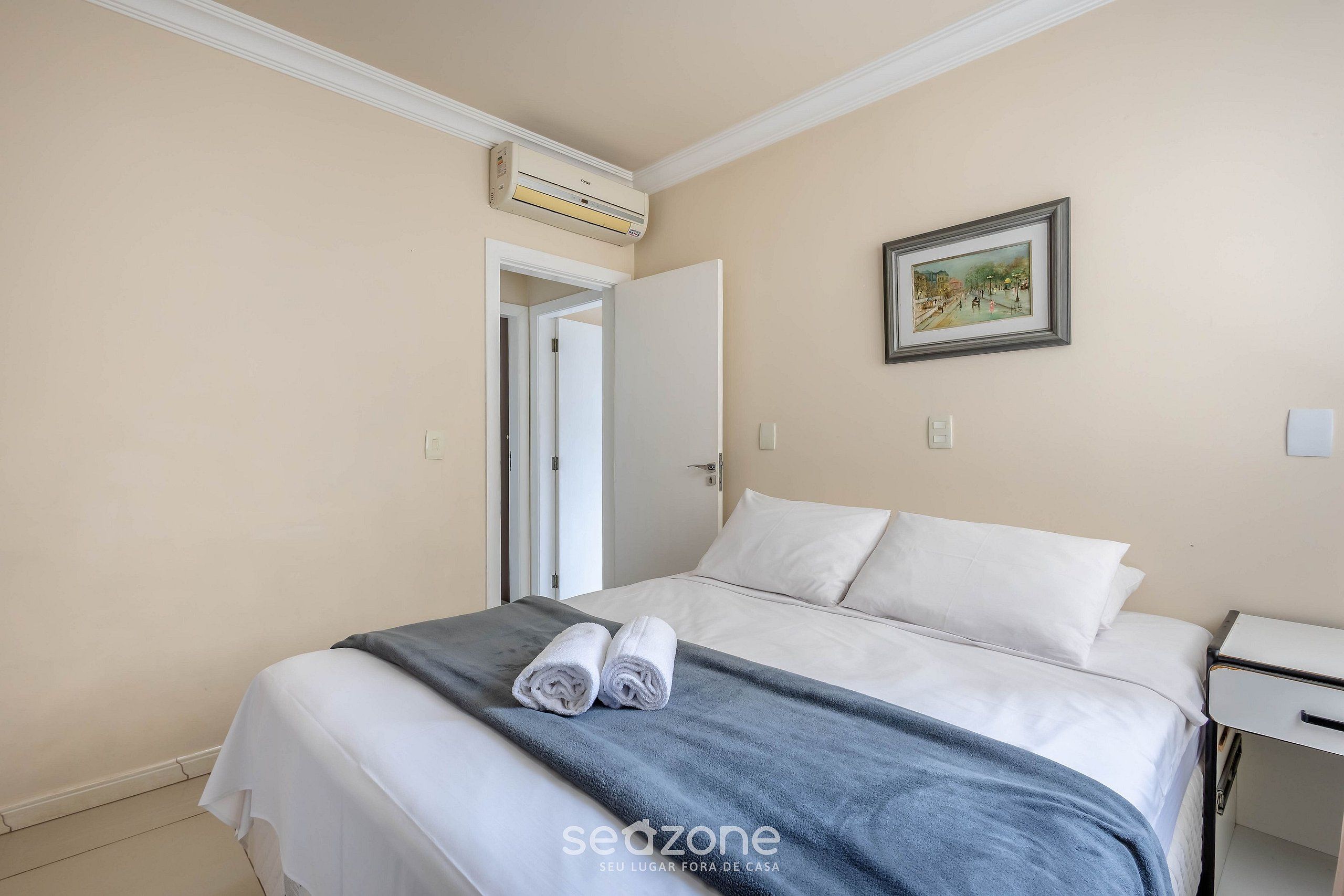 A hotel room with a bed, a bedside table, a wall-mounted air conditioner, and a framed picture on the wall.