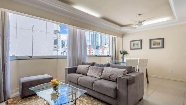 A modern living room with a grey sofa, a glass coffee table, and a view of tall buildings through large windows. - Apts Just 210m From the Beach in Balneario Camboriu - Edis (Balneário Camboriú)