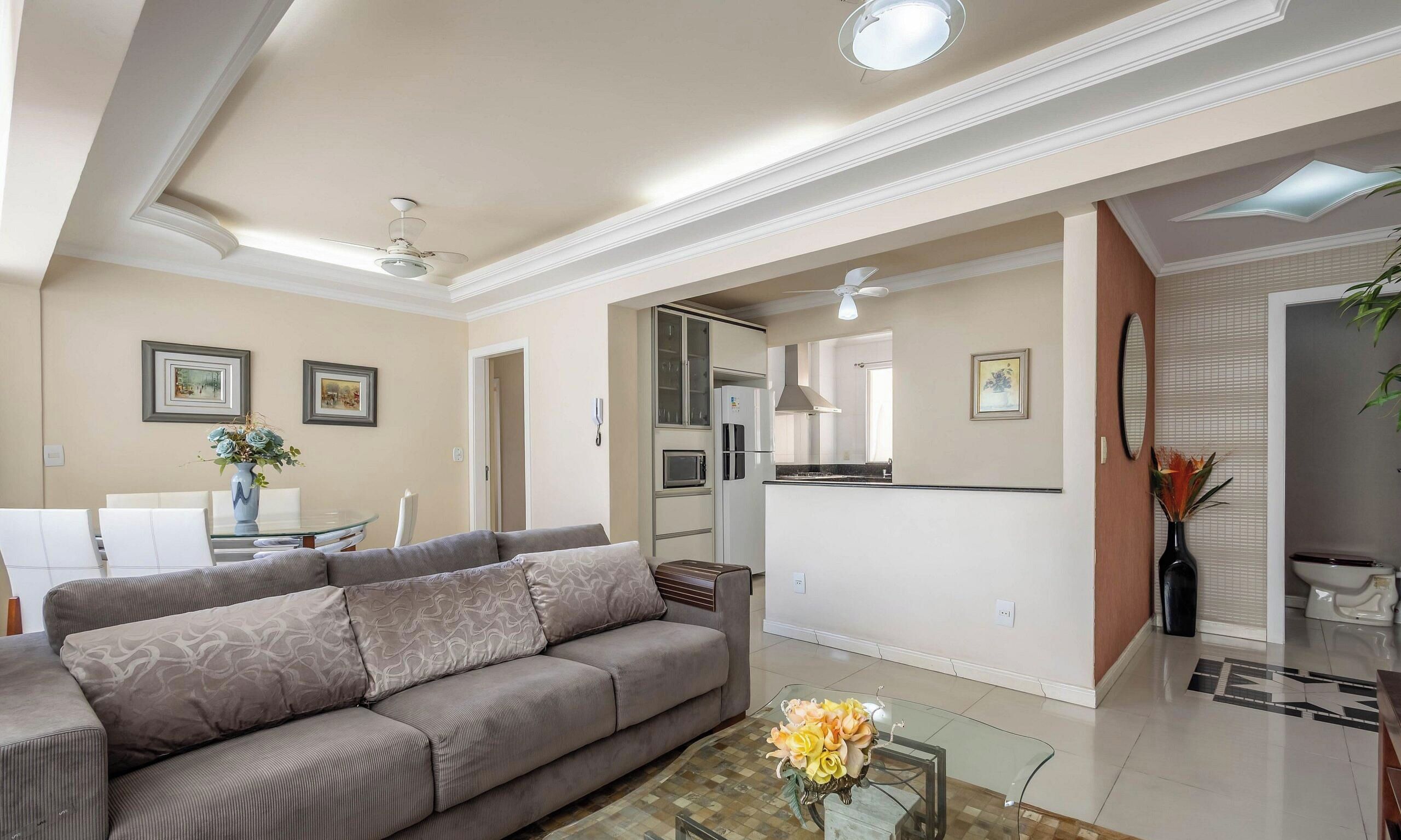 Uma sala de estar moderna com um sofá, uma mesa de centro de vidro e uma cozinha ao fundo.