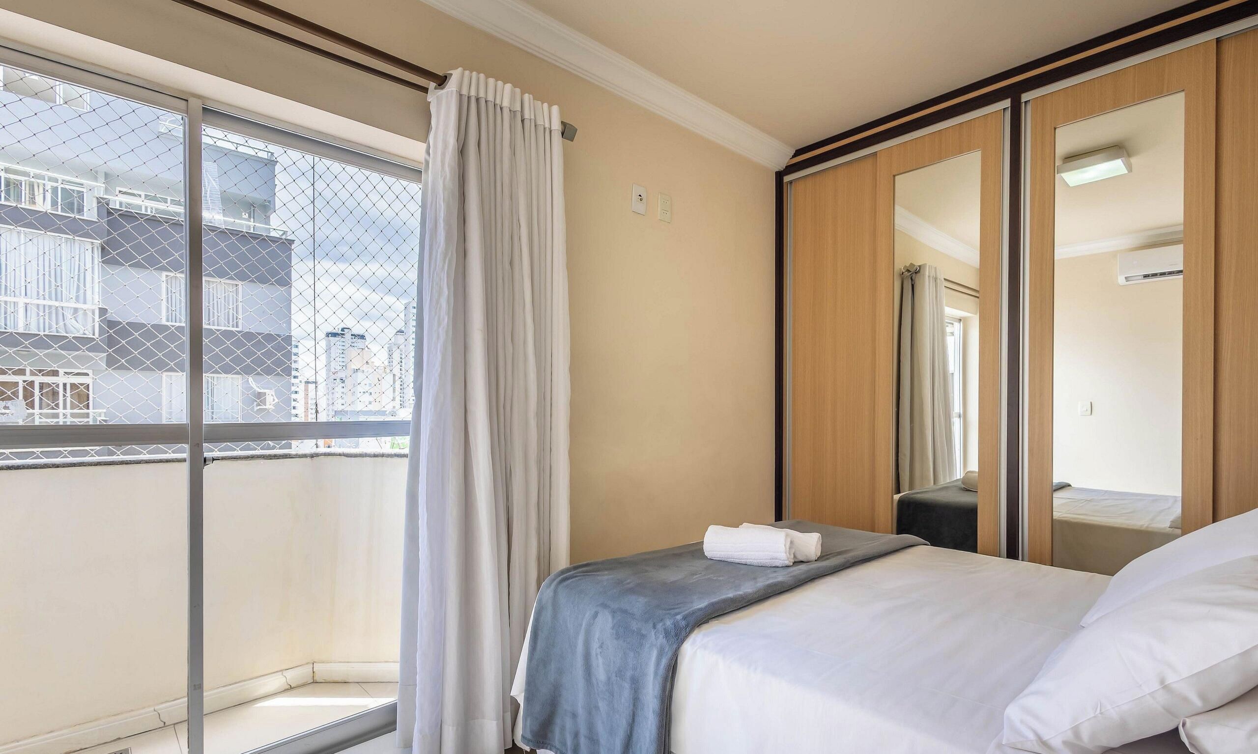A hotel room with a bed, a window with a view of buildings, a mirror, and a wardrobe.
