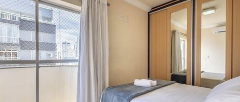 A hotel room with a bed, a window with a view of buildings, a mirror, and a wardrobe.
