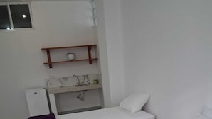 Una habitación con dos camas de madera, cada una con ropa de cama blanca y una manta morada. Hay un estante sobre un lavabo blanco con un espejo y una toalla.
