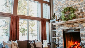 A cozy living room with a stone fireplace, large windows, and a ceiling fan.