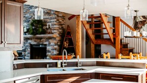 A modern kitchen with a stone fireplace, wooden staircase, and pendant lighting.