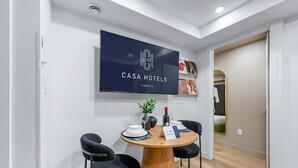Una habitación de hotel moderna con un televisor de pantalla plana mostrando el nombre del hotel, una mesa de comedor de madera con sillas y una planta pequeña.
