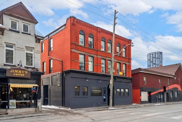 A red brick building with a black awning, a sign for 'The McCul' coffee shop, and a clear sky. - The McCaul | Basement Suite Near Chinatown (Toronto)
