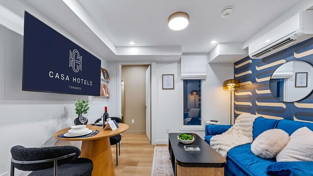 Una sala de estar moderna con un sofá azul, una mesa de comedor de madera y un televisor montado mostrando 'CASA HOTELS TORONTO'.