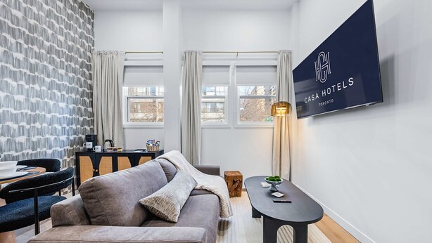 Moderni hotellihuone, jossa on harmaa sohva, pieni pöytä, seinälle asennettu televisio ja ikkuna, jossa on verhot.