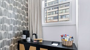 Hotellihuone, jossa on ikkuna, musta konsolipöytä, jossa on kahvinkeitin, pullovesipulloja sisältävä kori ja kuvioitu seinä.