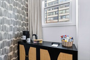 Una habitación de hotel con ventana, una mesa consola negra con cafetera, un cesto de agua embotellada y una pared con diseño decorativo.